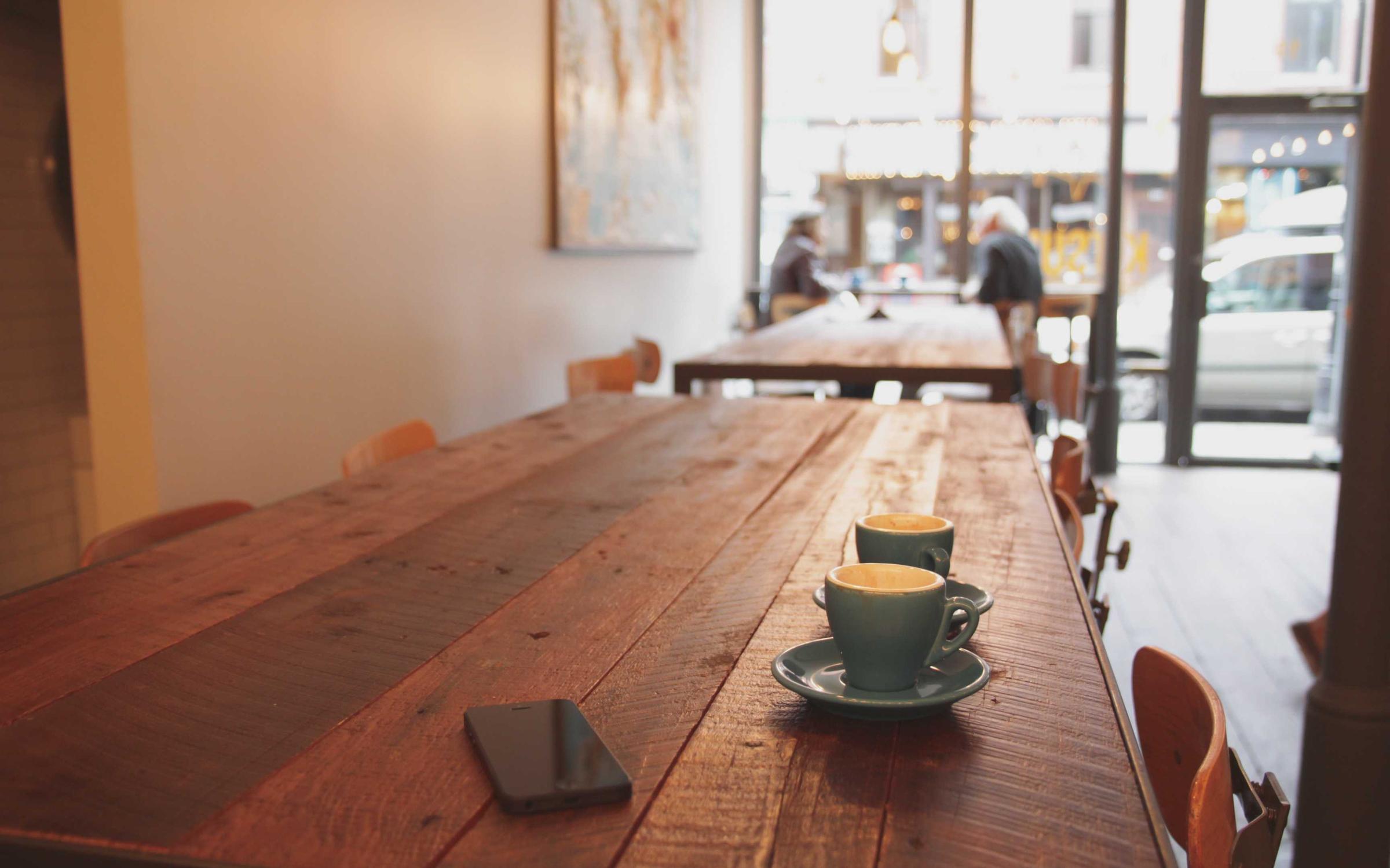 A long wooden table with two espresso cups, looking toward a bright street window