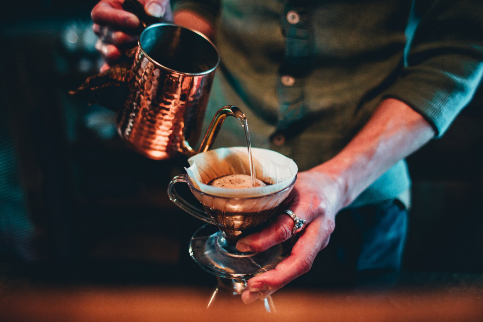 A barista pouring water from a copper kettle over a pour-over dripper